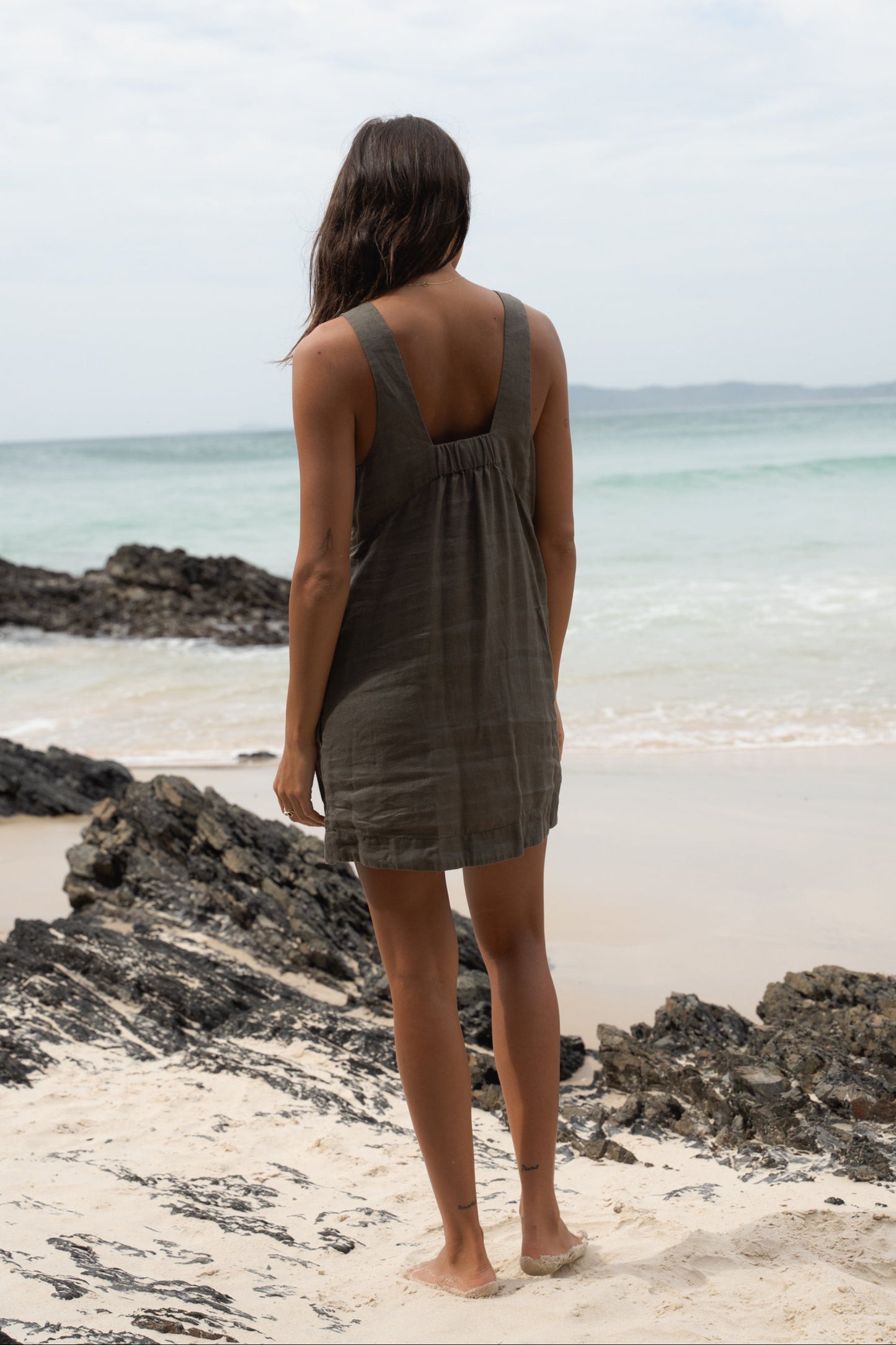 LILLY PILLY w woman standing on a beach wearing a Isla linen dress in Khaki showing back view