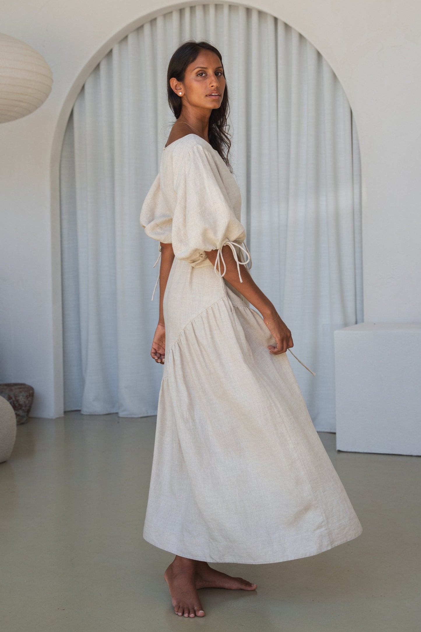 LILLY PILLY woman in an oatmeal Charlotte linen dress side view in a minimalistic room with a lamp and shelves.