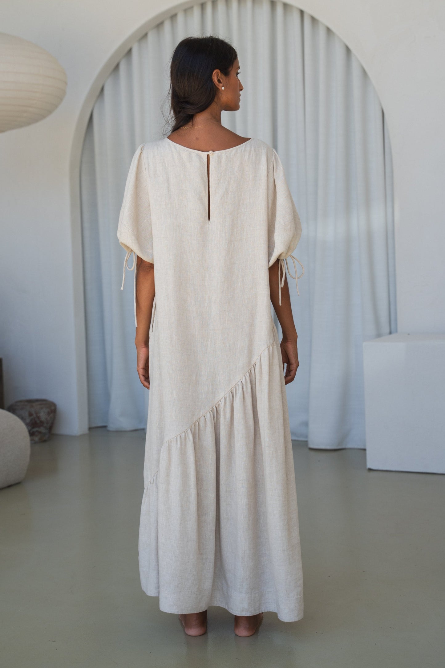 LILLY PILLY woman in an oatmeal Charlotte linen dress showing back view while standing in a minimalistic room with a lamp and shelves.