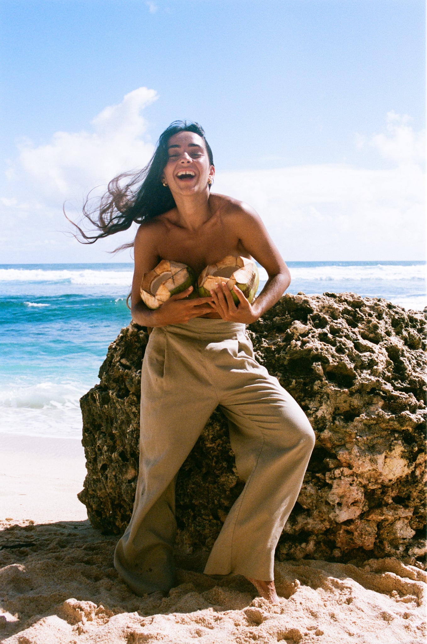 Person sitting on a rock by the ocean holding coconuts, wearing Ivy taupe linen pants with a clear blue sky.