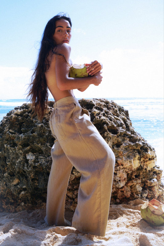 LILLY PILLY woman holding a coconut on a rocky beach in Ivy taupe linen pants with ocean in the background
